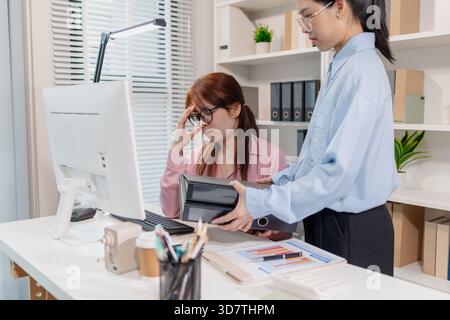 Two female colleagues working in an office, one looking stressed with her hand on her forehead while the other offers a folder, symbolizing teamwork a Stock Photo