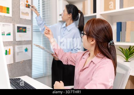 Two Asian office colleagues working together in the same department, reviewing documents and discussing project details during a team meeting, showing Stock Photo