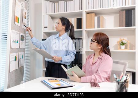 Two Asian office colleagues working together in the same department, reviewing documents and discussing project details during a team meeting, showing Stock Photo