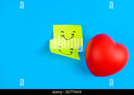 Red heart and two smiley sticky notes on blue background, symbolizing love, happiness, and emotional connection through vibrant contrast. Stock Photo