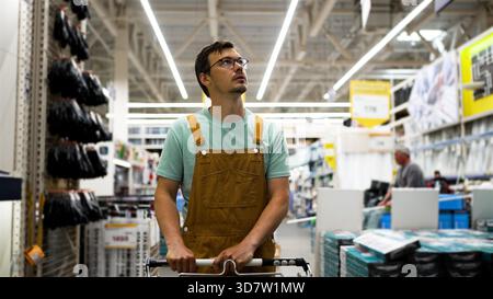 Young construction worker wearing brown overalls and glasses looking up while pushing a shopping cart in a large hardware store, searching for building materials Stock Photo