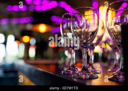 Empty clear wineglasses stand in rows on bar counter at nightclub, blurred purple neon and yellow lights on background. Clean cocktail glasses ready f Stock Photo