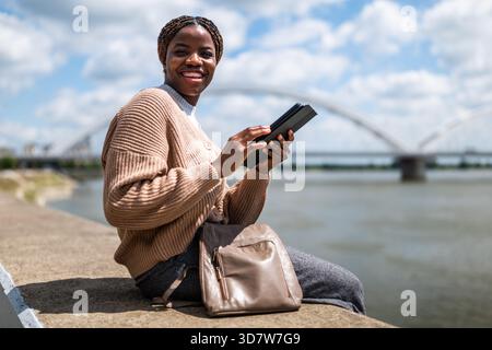 Young black woman is relaxing outdoor by the river. She is using digital tablet. Stock Photo