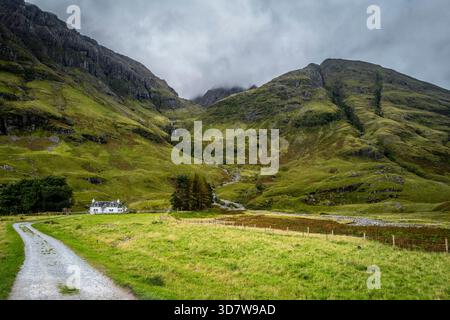 Serene landscape of a green valley with a white cottage and majestic mountains under a cloudy sky. Glencoe, Scotland Stock Photo