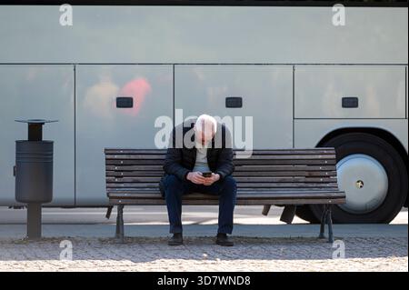 Silute, Lithuania - May 25, 2025: Elderly man focused on his smartphone while sitting alone on a bench at a bus station. Stock Photo