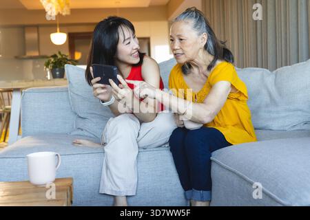 Pointing at smartphone Asian grandmother and granddaughter sitting on sofa at home, with coffee mug. Lifestyle, multigeneration, bonding, connection, comfort, warmth, contemporary || Model released Stock Photo