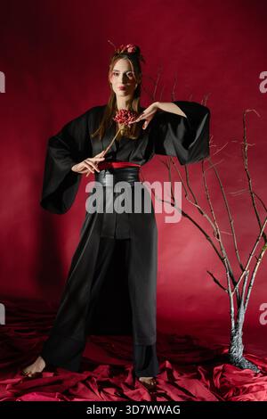 A woman stands gracefully in a black traditional Japanese dress within a studio. She holds a flower and poses against a striking red backdrop, creating a captivating visual. || Model released Stock Photo
