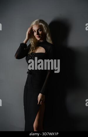 A woman stands gracefully with one hand in her hair, wearing a stylish black dress. The soft lighting creates shadows, enhancing the artistic atmosphere around her. || Model released Stock Photo