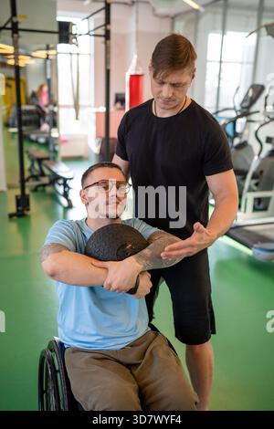 Disabled man in wheelchair doing exercise with heavy med ball under supervision of personal trainer. Rehabilitation coach guiding with movements, controls exercise technique for injured male athlete. || Model released Stock Photo