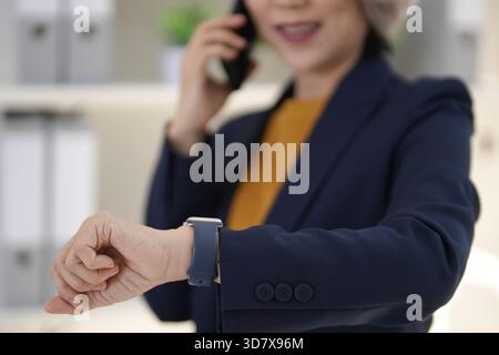 Mature Asian entrepreneur communicates using mobile technology checking wrist device professional setting productivity focus business efficiency Stock Photo