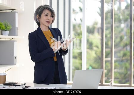 Asian older business woman diligently taking notes managing tasks and developing strategy in a contemporary office setting with a laptop. Stock Photo