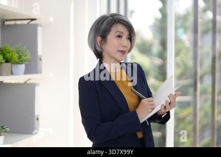 Mature Asian female professional thoughtfully captures ideas in a notebook within a bright contemporary workspace environment Stock Photo