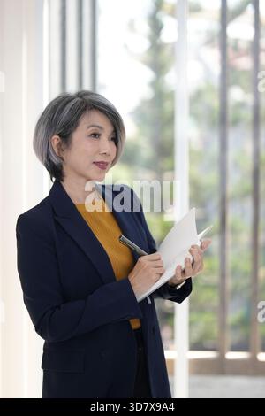 An elegant Asian senior businesswoman stands bright office writing notes. Plans future business strategies pen notebook professionalism. Stock Photo