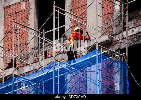 A construction worker wearing safety gear works on scaffolding at an urban building site, focusing on masonry and structural repair. Stock Photo
