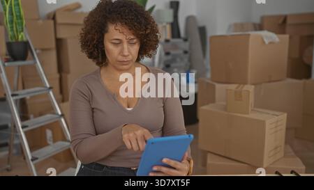 Hispanic woman tapping tablet while seated at table in building surrounded by moving boxes and packing materials; serenity. Stock Photo