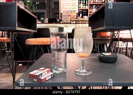 Two used beer glasses, cigarette packs, and an ashtray sit on a bar table in a dimly lit pub interior. Stock Photo