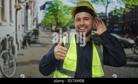 Man builder in yellow hardhat and high visibility vest smiling and waving right hand on street with parked cars and bicycle nearby; pride safety. Stock Photo