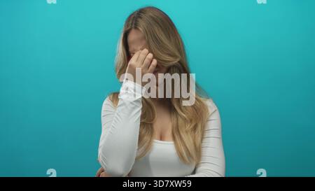 Young caucasian blonde woman pinches nose bridge under studio lights; stress headache fatigue tension. Stock Photo