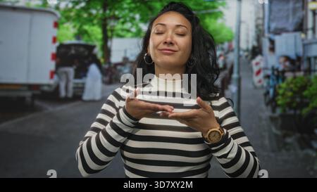 Woman blowing a kiss with hands visible on a city street wearing a black and white striped turtleneck and wooden watch; playful affection. Stock Photo