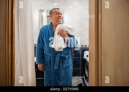 Adult caucasian happy man in blue bathrobe with white towel leaves bathroom. Daily personal care routine, self-care. Morning Shaving Routine Stock Photo