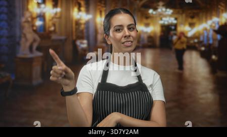 Young brunette woman wearing a striped apron points finger to object while standing in ornate museum building lobby; welcoming hospitality. Stock Photo