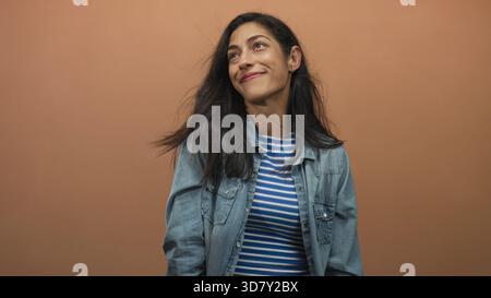 Woman smiling with hair blowing, wearing denim jacket and blue striped crop top in peach studio; contentment. Stock Photo