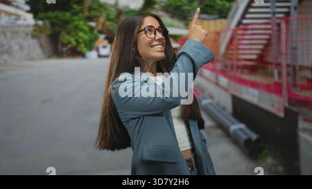 Woman in blue blazer wearing glasses points finger upward on a street beside construction fencing and pipes; joyful curiosity. Stock Photo