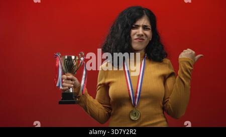 Young arab brunette woman holds trophy and points thumb over shoulder in studio with red backdrop; reluctant pride. Stock Photo