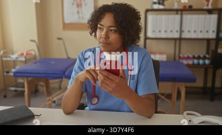 Woman in blue scrubs wearing stethoscope sits at desk taps red smartphone in clinic room; professional focus calm determination. Stock Photo