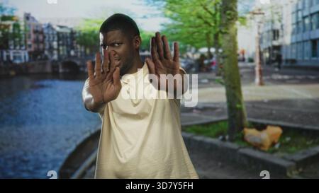 Man holding palms forward in stop gesture on street canal walkway by water, facing camera with guarded expression; rejection. Stock Photo
