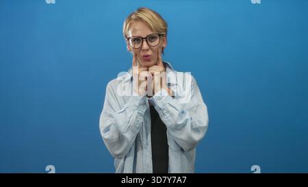 Caucasian blond woman in glasses pressing index fingers to her cheeks while puffing cheeks in a blue studio; playful mood. Stock Photo