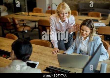 Two women discuss ideas while working on a laptop in a contemporary office setting. The atmosphere is focused and collaborative, promoting teamwork an Stock Photo