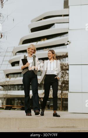 Two women are walking together in a contemporary city setting. They are dressed in professional attire, smiling and engaging in conversation, showcasi Stock Photo