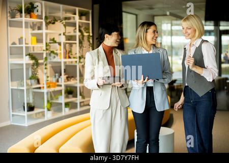 In a well-lit office, three women in professional attire share ideas and laugh while holding a laptop and a tablet. Plants and modern decor enhance th Stock Photo