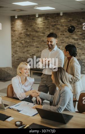 Four professionals gather around a table in a well-lit office, smiling and discussing ideas. The atmosphere is friendly, with one person shaking hands Stock Photo