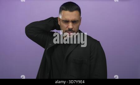 Young hispanic man wearing black shirt touches neck in purple studio with eyes closed and tense expression; discomfort. Stock Photo