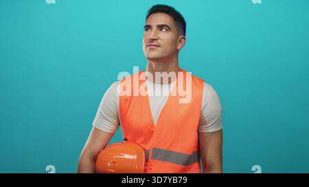 Man holding orange hard hat and wearing orange safety vest in studio against turquoise backdrop; safety professionalism optimism. Stock Photo