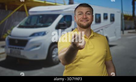 Man in yellow polo beckoning with fingers on street beside white motorhome campervan, smiling and leaning forward toward camera; inviting travel adven Stock Photo