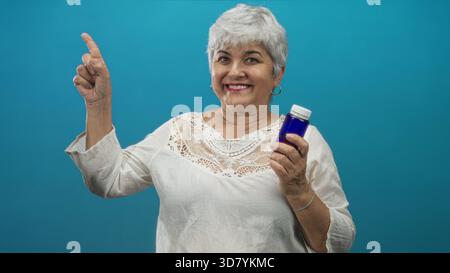 Senior woman with grey hair and plus size holding a blue medicine bottle points finger and smiles in studio; wellness confidence. Stock Photo