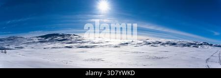 Vast panoramic winter landscape showing rolling snow covered hills and mountains under a brilliant sun and blue sky, with visible tracks on the pristi Stock Photo