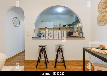 Kitchen with arched opening, tile backsplash, bar stools, and compact dining setup. Stock Photo