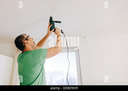 Man using electric drill to make hole in white ceiling at home. Stock Photo