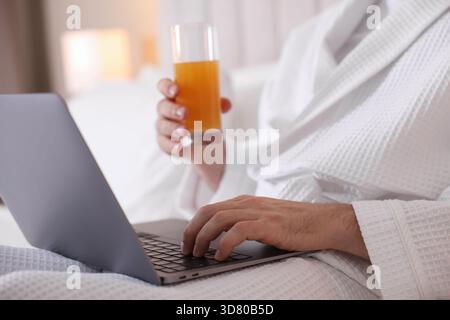 Man in bathrobe with glass of juice working on laptop indoors, closeup Stock Photo