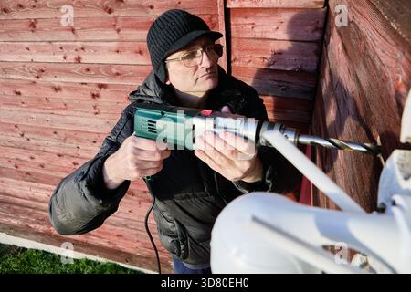 Creating bore for security camera wiring, technician operates electric drill on exterior wall of country home, installer makes hole for cable route. Stock Photo