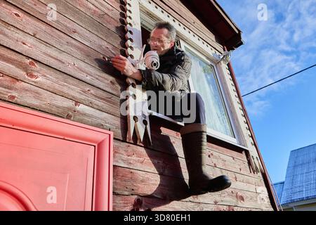 Pulling surveillance camera cable through drilled passage in wall, installer sits on window frame and feeds wire toward exterior mounting point. Stock Photo