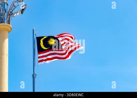 Malaysian national flag waving on a metal pole near a yellow column against a clear blue sky Stock Photo