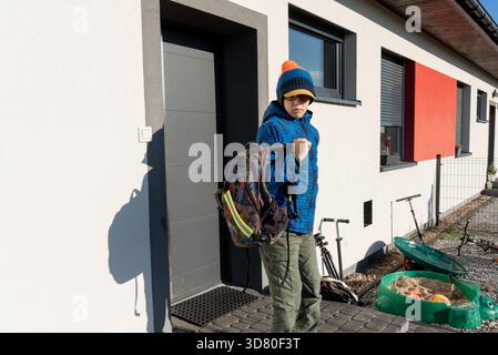 Boy in blue winter jacket and sunglasses putting on backpack near modern house entrance on sunny morning. Concept of going to school, childhood indepe Stock Photo