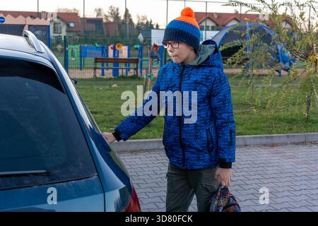 Boy in blue winter jacket and glasses holding bag while touching car near playground in suburban area on autumn day. Concept of after school pickup, c Stock Photo