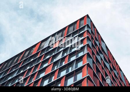 Low-angle view of a modern office building with a striking red and grey facade and repetitive window pattern, set against a bright, cloudy sky. Stock Photo