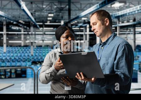 Data center computer scientists doing brainstorming, using laptop to manage system stability. Server room colleagues optimizing performance with device, configuring equipment Stock Photo